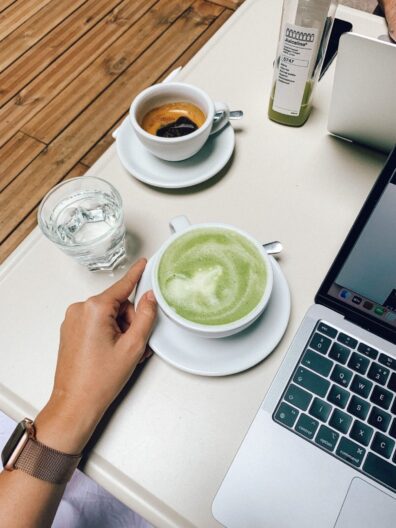 A cup of matcha latte, espresso and a glass of water on a table next to a laptop